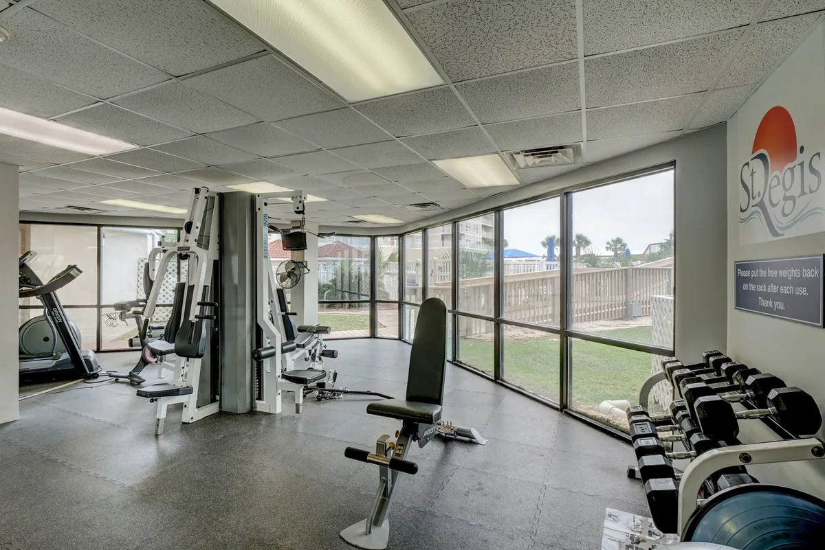 A bright and spacious fitness center equipped with various gym equipment, including cardio and strength machines. Large windows let in natural light and offer views of the outdoors, while exercise mats on the floor provide space for stretching or group fitness classes.