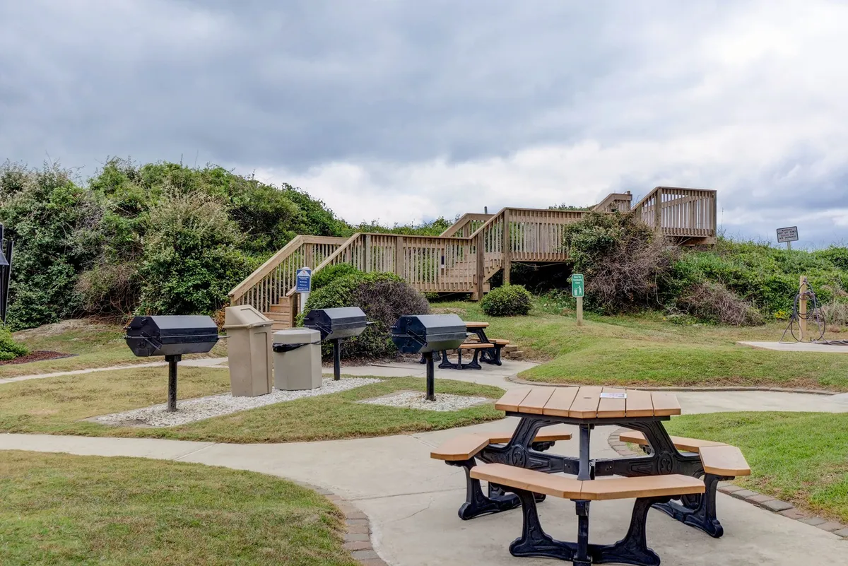 An outdoor area featuring a picnic table and multiple grill stations, with a staircase leading up. The setting conveys a relaxed mood, perfect for barbecuing, picnicking, and social gatherings under the open sky.