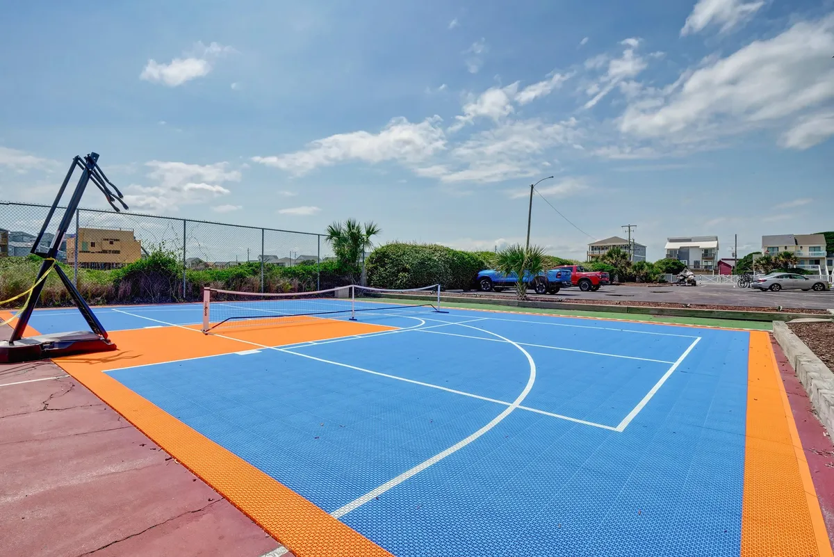 A vibrant, multi-purpose outdoor court featuring a basketball hoop, a tennis net (suitable for pickleball as well), and a colorful playing surface. The area invites both competitive and casual games under open skies, surrounded by green landscaping.