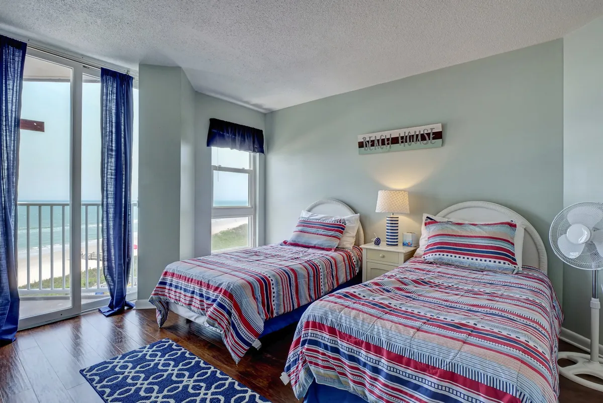 A cozy guest bedroom featuring two twin beds adorned with soft bedding, large windows dressed in blue curtains, and a nightstand with a lamp between the beds. The windows allow natural light to pour in, with a hint of coastal scenery visible, reinforcing the beachside setting.