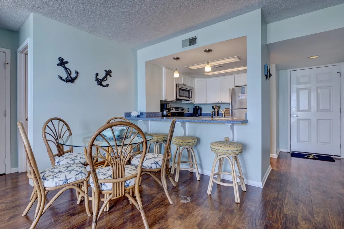 A bright dining area with a glass dining table surrounded by rattan chairs, adjacent to a kitchen counter. Nautical anchor decor and plenty of natural light create a relaxed and inviting setting, perfect for family meals or casual gatherings.