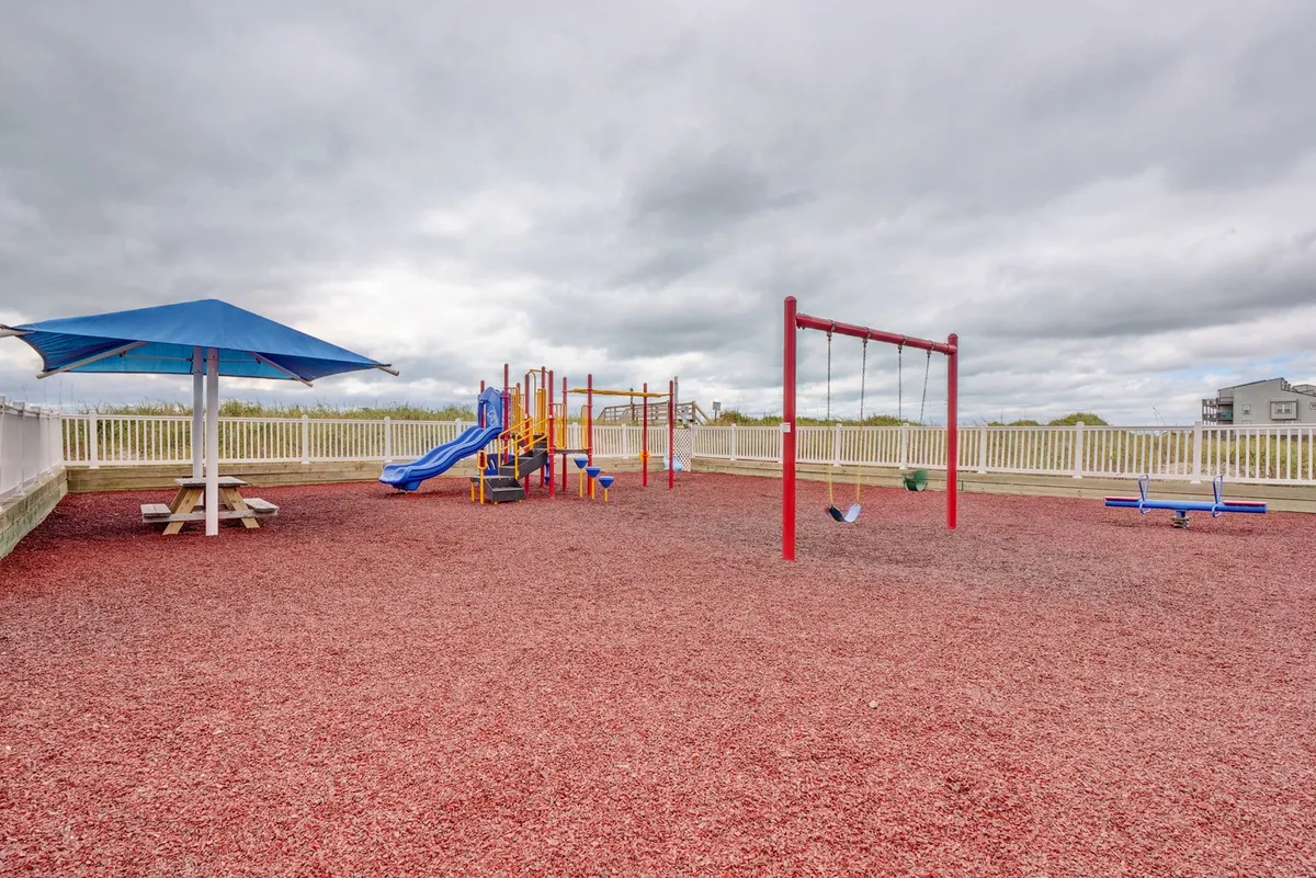 This image features a vibrant outdoor playground complete with swings, playground equipment, and a shaded picnic table under an umbrella. The setting is lively, inviting, and perfect for children to play while families gather nearby.