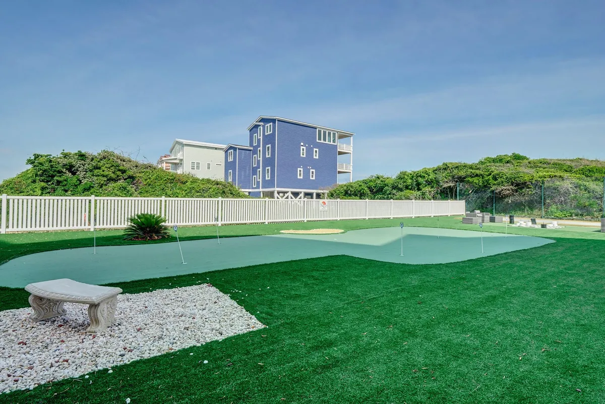 A relaxing outdoor scene with a green putting green, a stone bench, a fenced yard, and a blue house in the background, offering opportunities for relaxation or outdoor gatherings after a productive day.