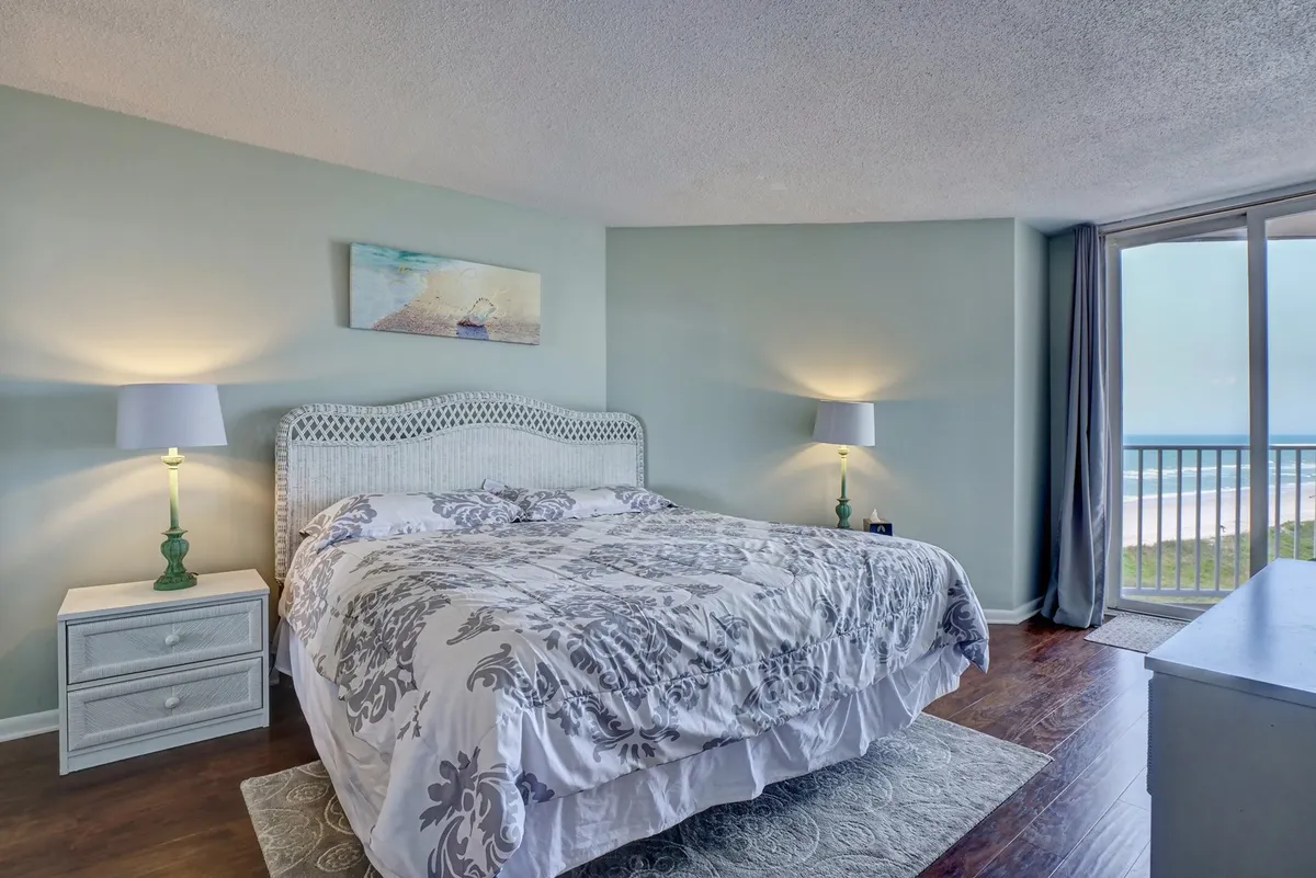 A serene bedroom with a plush bed positioned beside a large window that reveals expansive ocean views. Nightstands with lamps and a tasteful decorative painting add to the calming ambiance, creating a restful retreat. Soft natural light filters through, highlighting the tranquil setting.