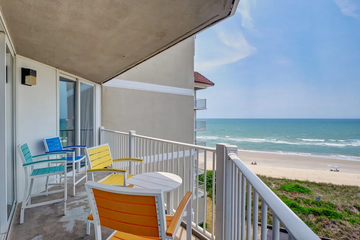 A tranquil exterior shot of a private covered balcony featuring colorful chairs and a small table, all looking out over an unobstructed ocean view. This inviting outdoor oasis is ideal for sipping morning coffee or enjoying sunset cocktails in a serene setting.