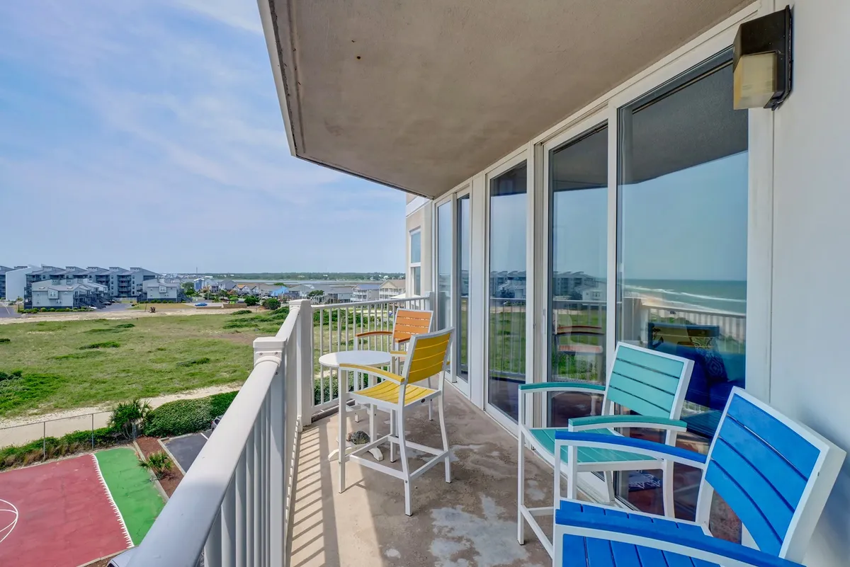 A cheerful exterior shot of a balcony with colorful chairs, a view of the landscape, and a glass railing. The scene suggests relaxation, outdoor socializing, and enjoying beautiful mornings and sunsets as a family.