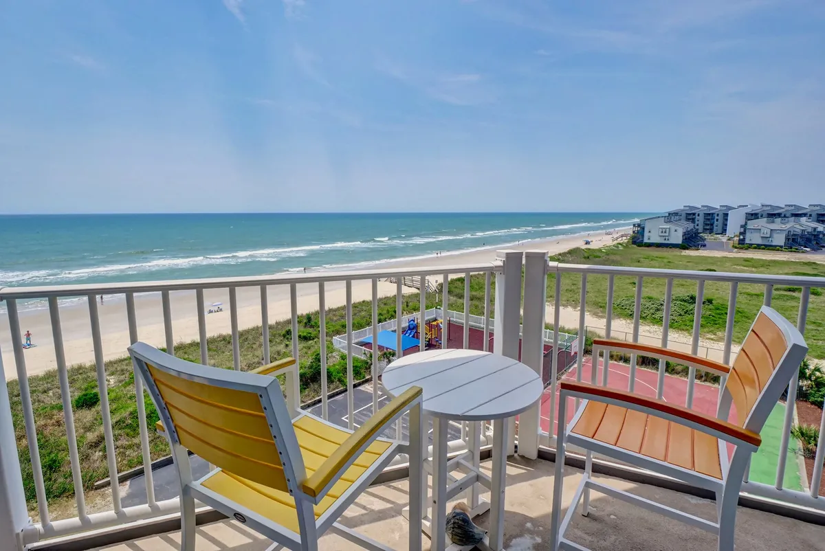 The image showcases a serene balcony scene with comfortable chairs overlooking a sweeping ocean view at North Topsail Beach. The sandy shoreline stretches alongside the turquoise waves beneath a vast, open sky. The setting is perfect for enjoying morning coffee, reading a book, or simply relaxing and soaking in the coastal beauty.