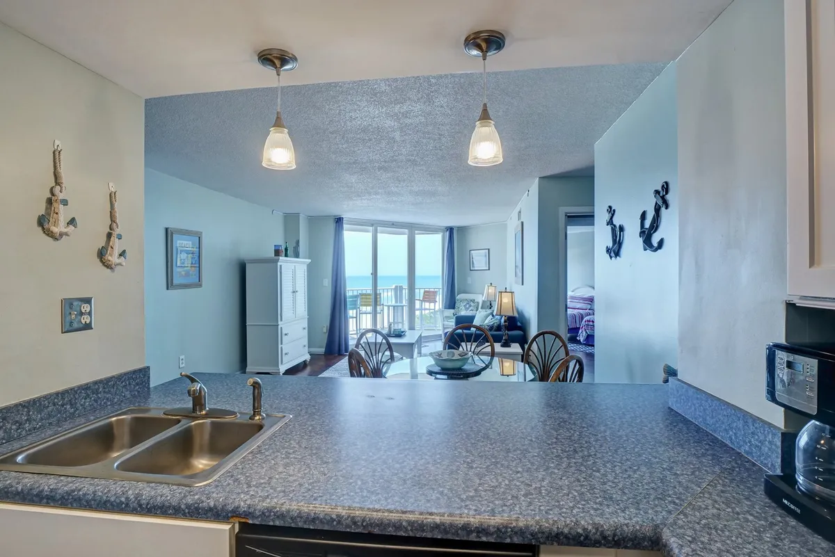 This inviting interior scene features a modern kitchen with stainless steel appliances, a dining area, and large windows framing a stunning ocean view. The space is flooded with natural light and designed for both meal preparation and relaxed dining with a scenic backdrop.