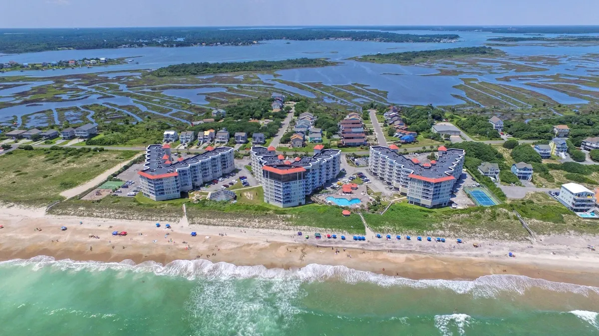 A vibrant coastal view featuring beachfront properties, a swimming pool, ocean waves, colorful umbrellas, and natural marshland. The scene is lively, yet tranquil, suggesting a blend of relaxation and opportunity for family fun or exploration.