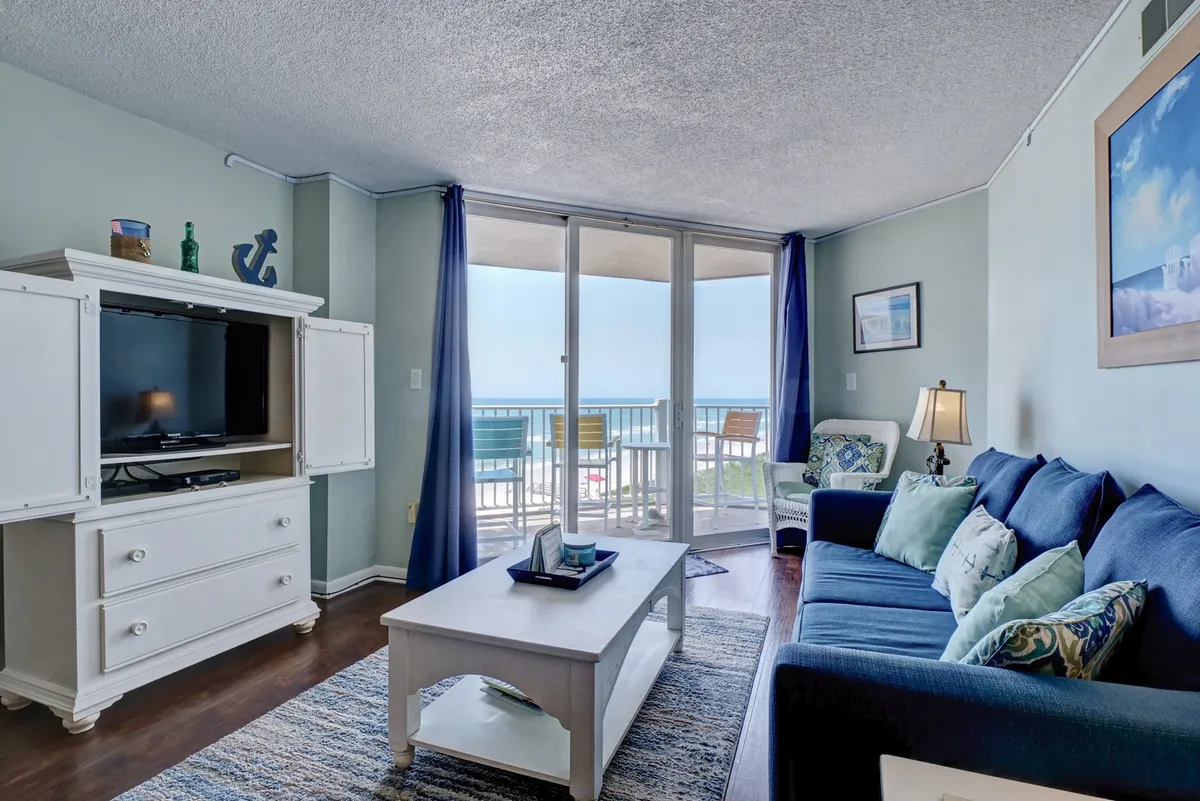 This image captures a cozy living room arranged around a plush sofa and coffee table, with a smart TV positioned for easy viewing and expansive windows offering a sweeping view of the ocean. The natural light fills the space, and a glimpse of the adjacent balcony invites relaxation.